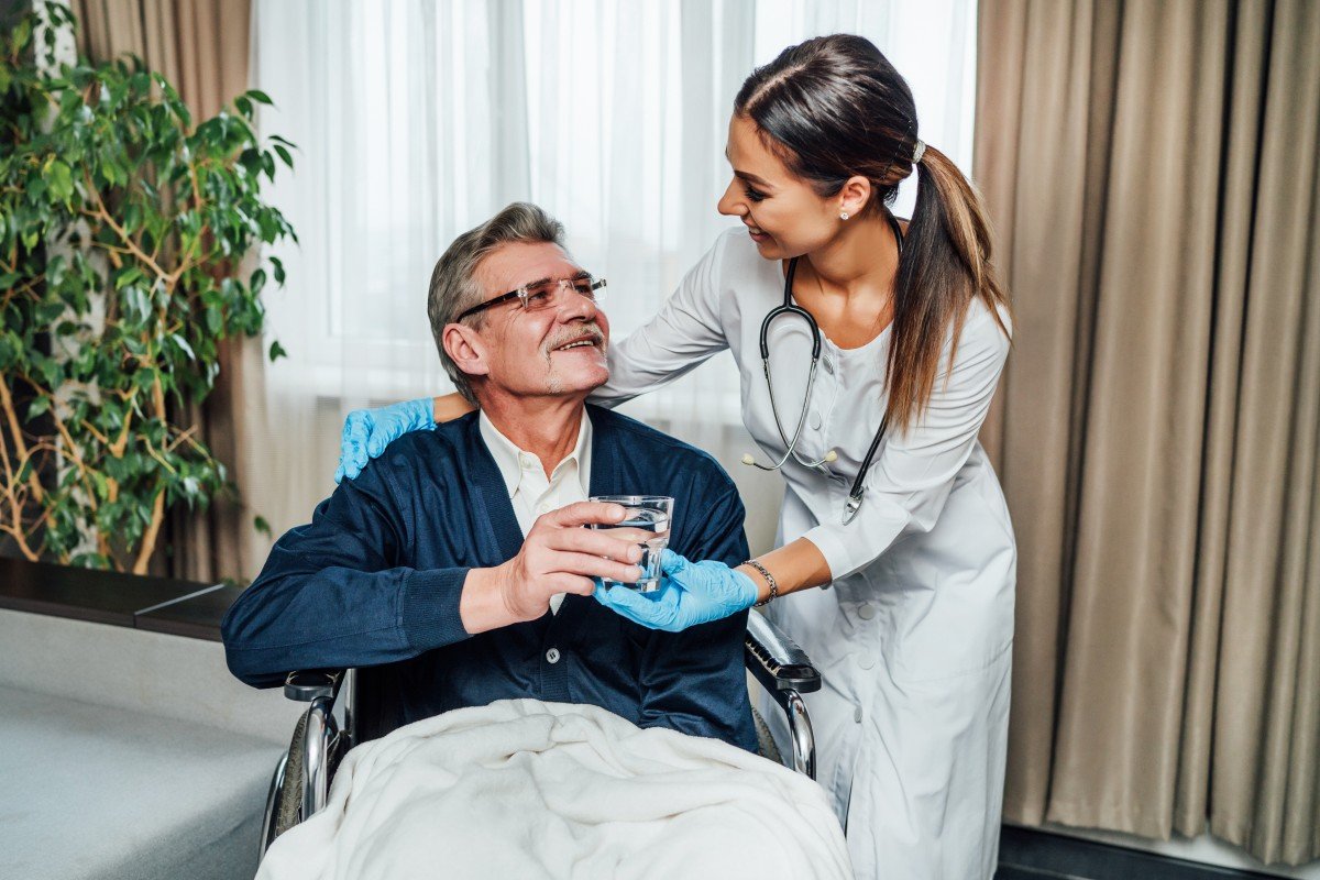 older man wheelchair smiles nurse assistant she hands him glass water (1)